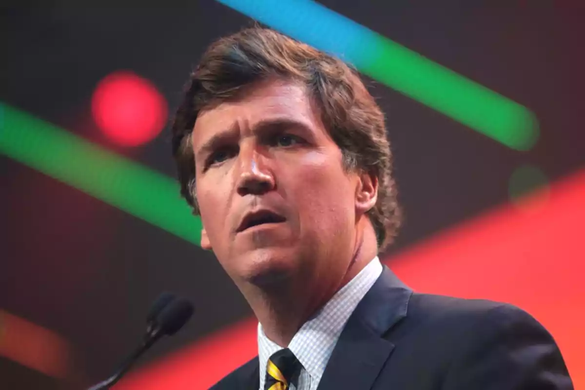 Brown-haired man in a suit and tie speaking in front of a microphone with blurred colored lights in the background Brown-haired man in a suit and tie speaking in front of a microphone with blurred colored lights in the background