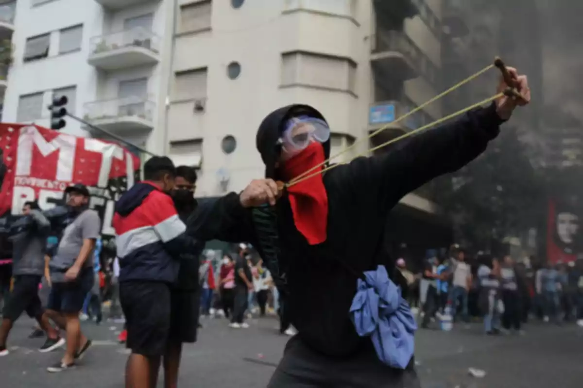 A person with a covered face and protective goggles uses a slingshot at a demonstration on an urban street with a building in the background and a group of people around.