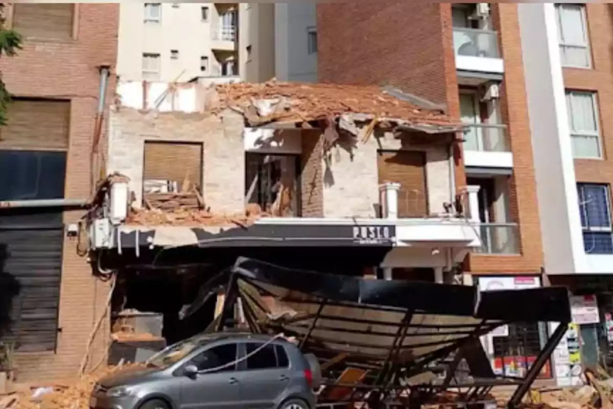 Edificio de dos pisos con la fachada parcialmente derrumbada, escombros sobre un auto gris estacionado y un toldo negro caído frente al local Edificio de dos pisos con la fachada parcialmente derrumbada, escombros sobre un auto gris estacionado y un toldo negro caído frente al local