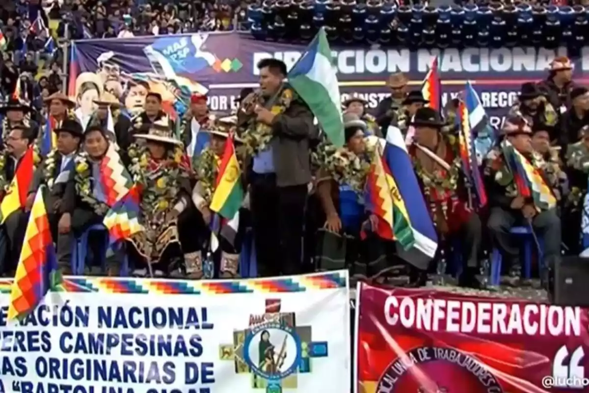 Un grupo de personas con banderas y guirnaldas se encuentra en un evento al aire libre, con pancartas que mencionan organizaciones campesinas y una multitud al fondo.