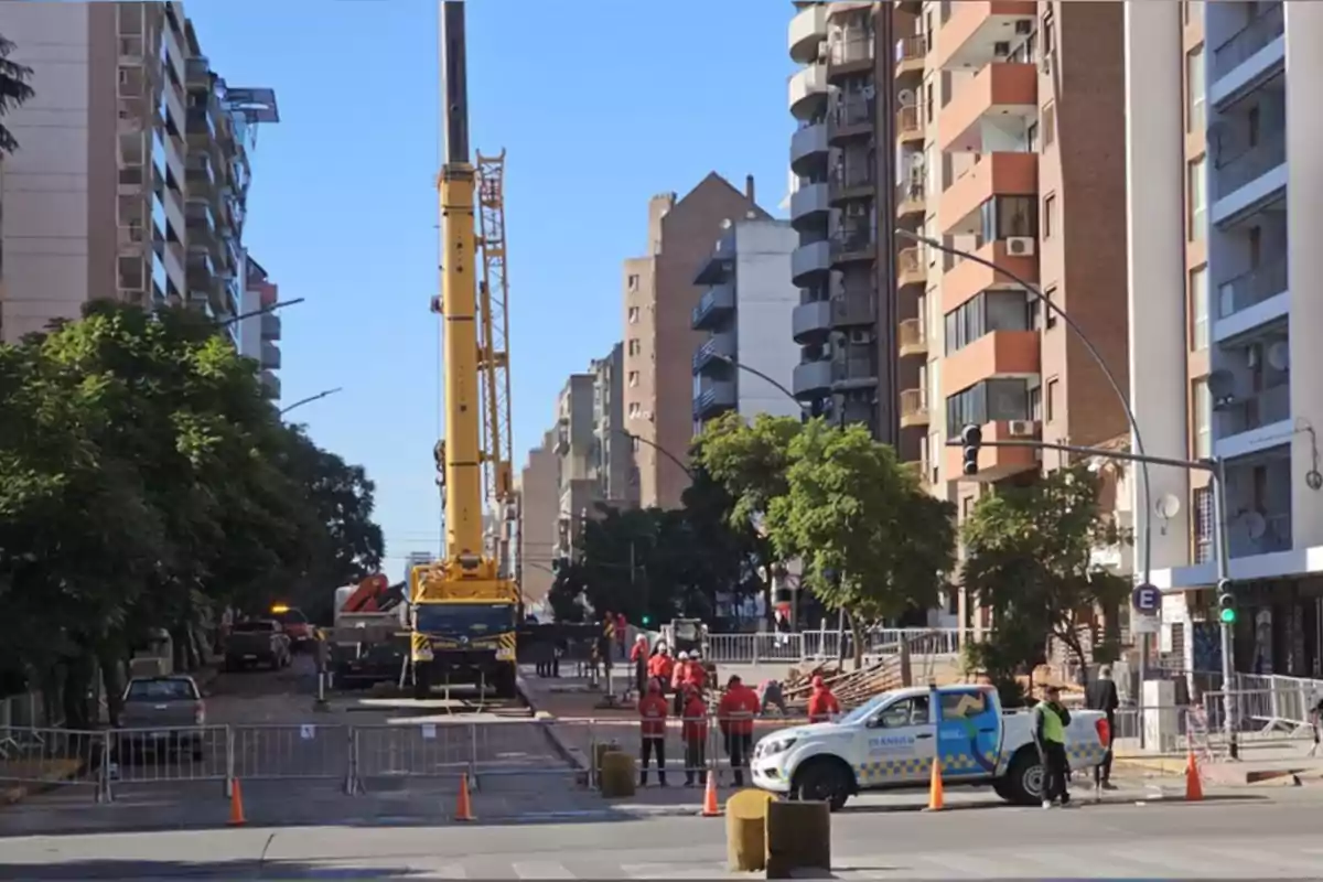 Una grúa amarilla de gran tamaño trabaja en una calle cerrada al tránsito en una zona urbana rodeada de edificios altos mientras varias personas con chalecos rojos y un vehículo de control de tránsito están presentes Una grúa amarilla de gran tamaño trabaja en una calle cerrada al tránsito en una zona urbana rodeada de edificios altos mientras varias personas con chalecos rojos y un vehículo de control de tránsito están presentes