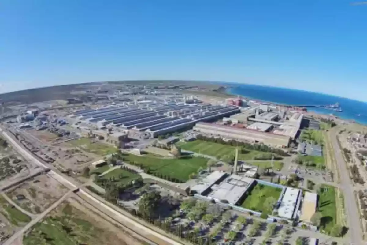 Vista aérea de un complejo industrial junto al mar con edificios, áreas verdes y un muelle. Vista aérea de un complejo industrial junto al mar con edificios, áreas verdes y un muelle.
