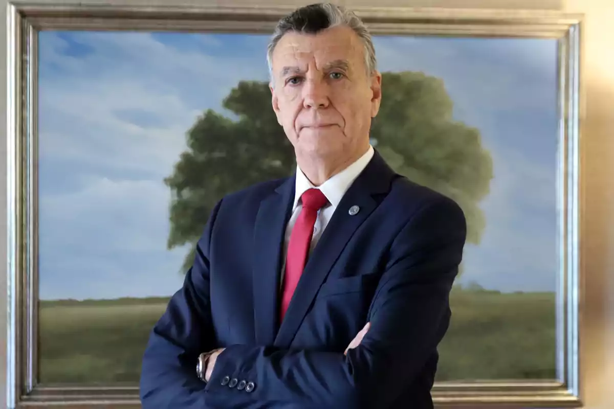 Un hombre de traje oscuro y corbata roja con los brazos cruzados frente a un cuadro de un paisaje con un árbol.