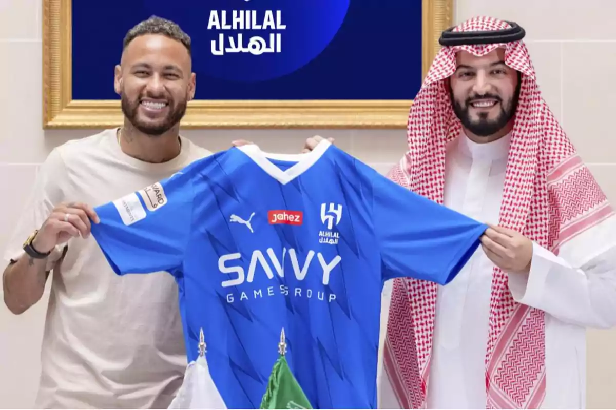 Dos personas sonrientes sostienen una camiseta azul del equipo Al Hilal con logotipos de patrocinadores.