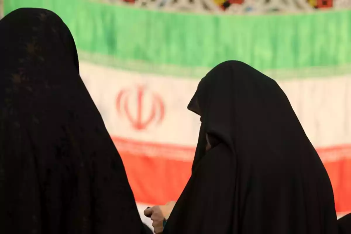 Dos mujeres con chador negro de espaldas frente a una bandera de Irán desenfocada