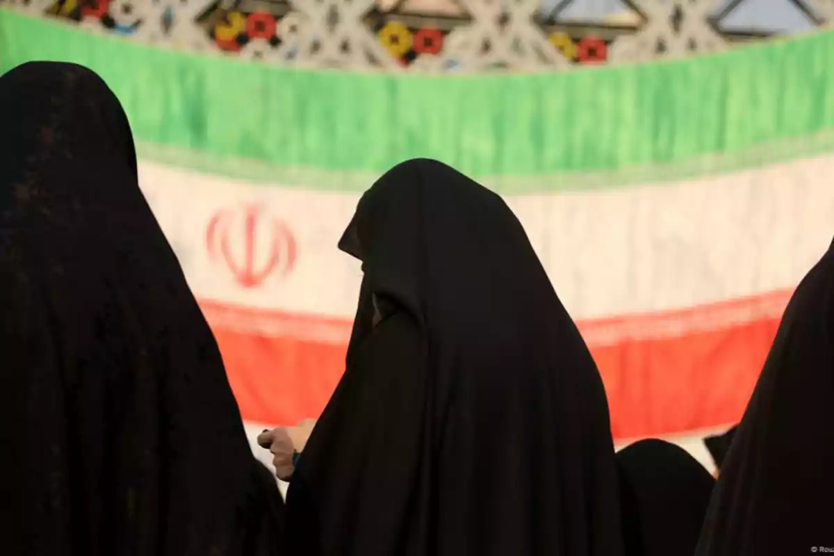 Personas vestidas con chador negro frente a una bandera de Irán.