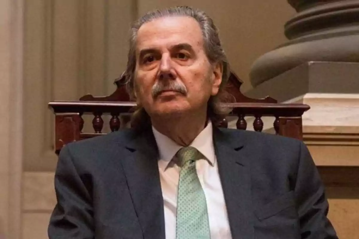 Gray-haired man with a mustache sitting on a wooden chair, wearing a dark suit and light green tie in a formal setting