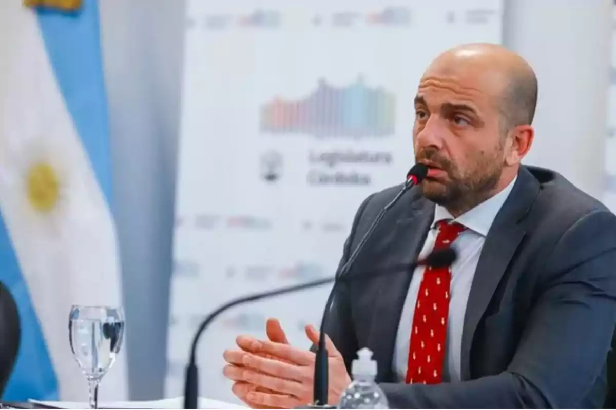 Un hombre con traje y corbata roja habla en un micrófono durante una conferencia de prensa con una bandera argentina de fondo.