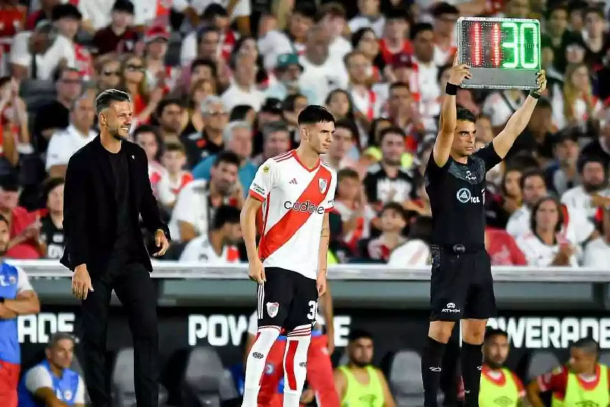 Un árbitro asistente sostiene un cartel de cambio mientras un jugador de River Plate se prepara para ingresar al campo y el entrenador observa desde la línea de banda con el estadio lleno de aficionados de fondo Un árbitro asistente sostiene un cartel de cambio mientras un jugador de River Plate se prepara para ingresar al campo y el entrenador observa desde la línea de banda con el estadio lleno de aficionados de fondo
