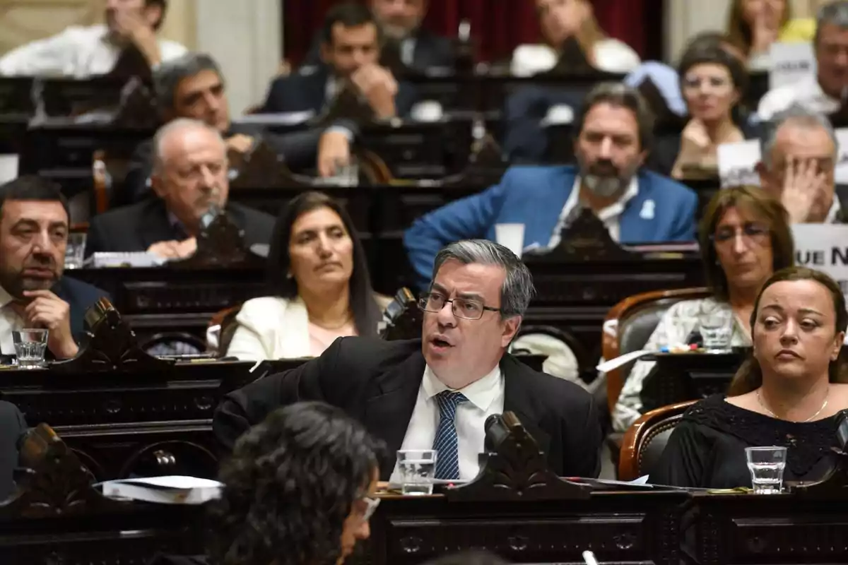 A group of people in a formal setting, possibly a parliament or congress, with a man in the foreground speaking while the others listen.
