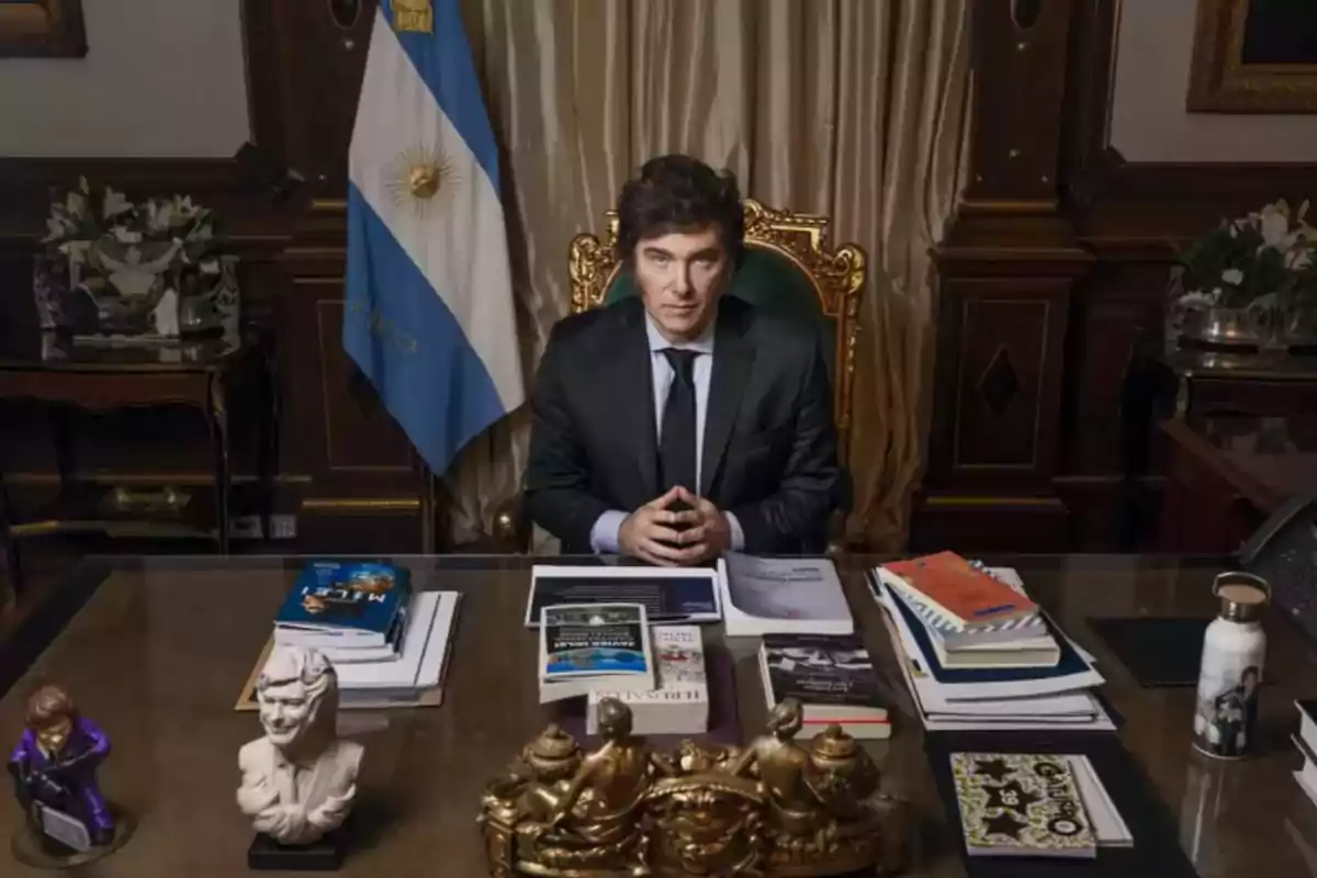 Un hombre sentado en un escritorio elegante con libros y adornos, frente a una bandera de Argentina.