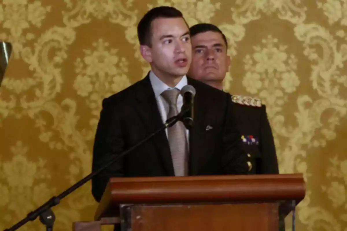 A man in a suit speaks at a podium with a microphone while another person in uniform stands behind him, with a decorative wall background.