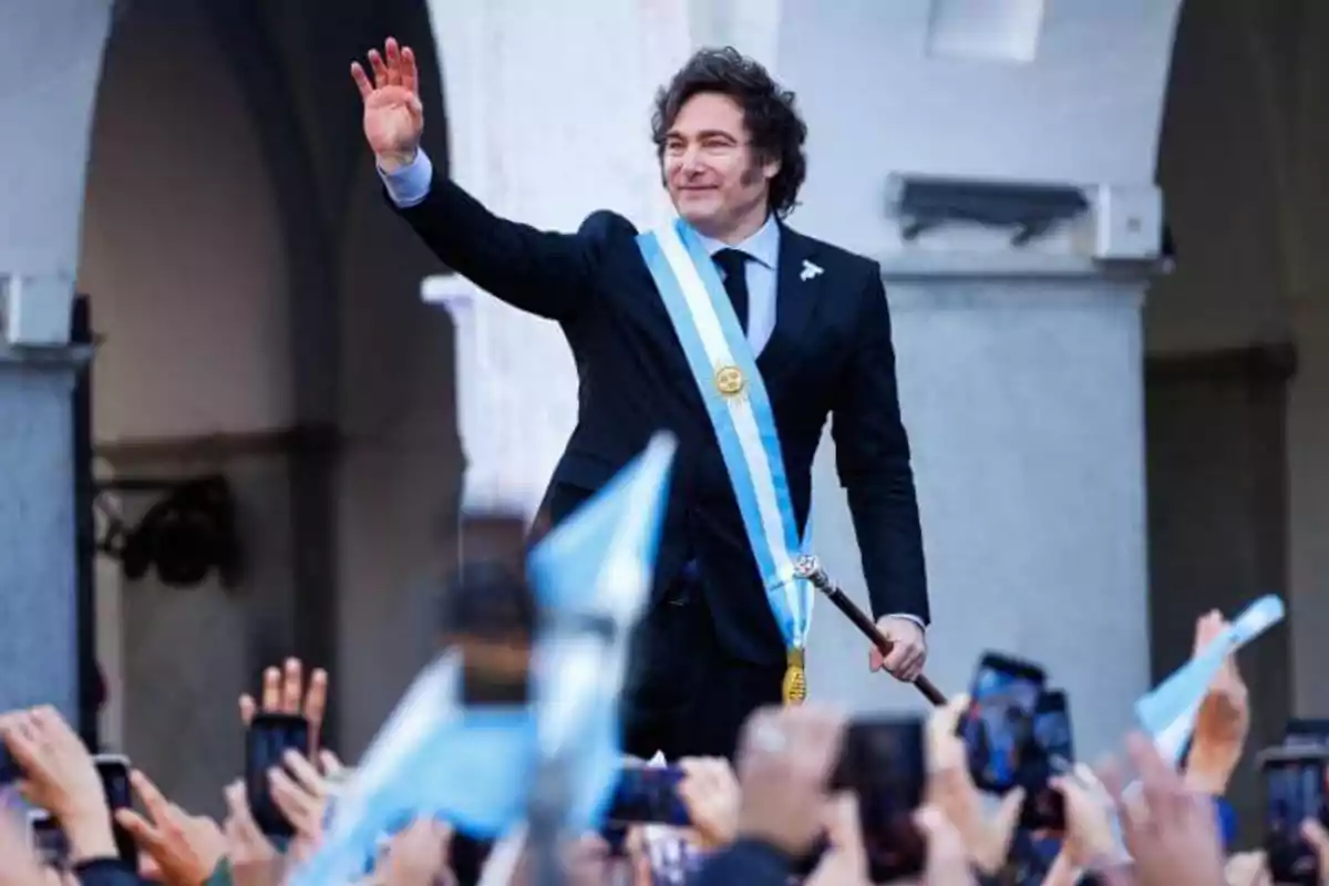 Un hombre con traje y banda presidencial saluda a una multitud que lo fotografía y ondea banderas.