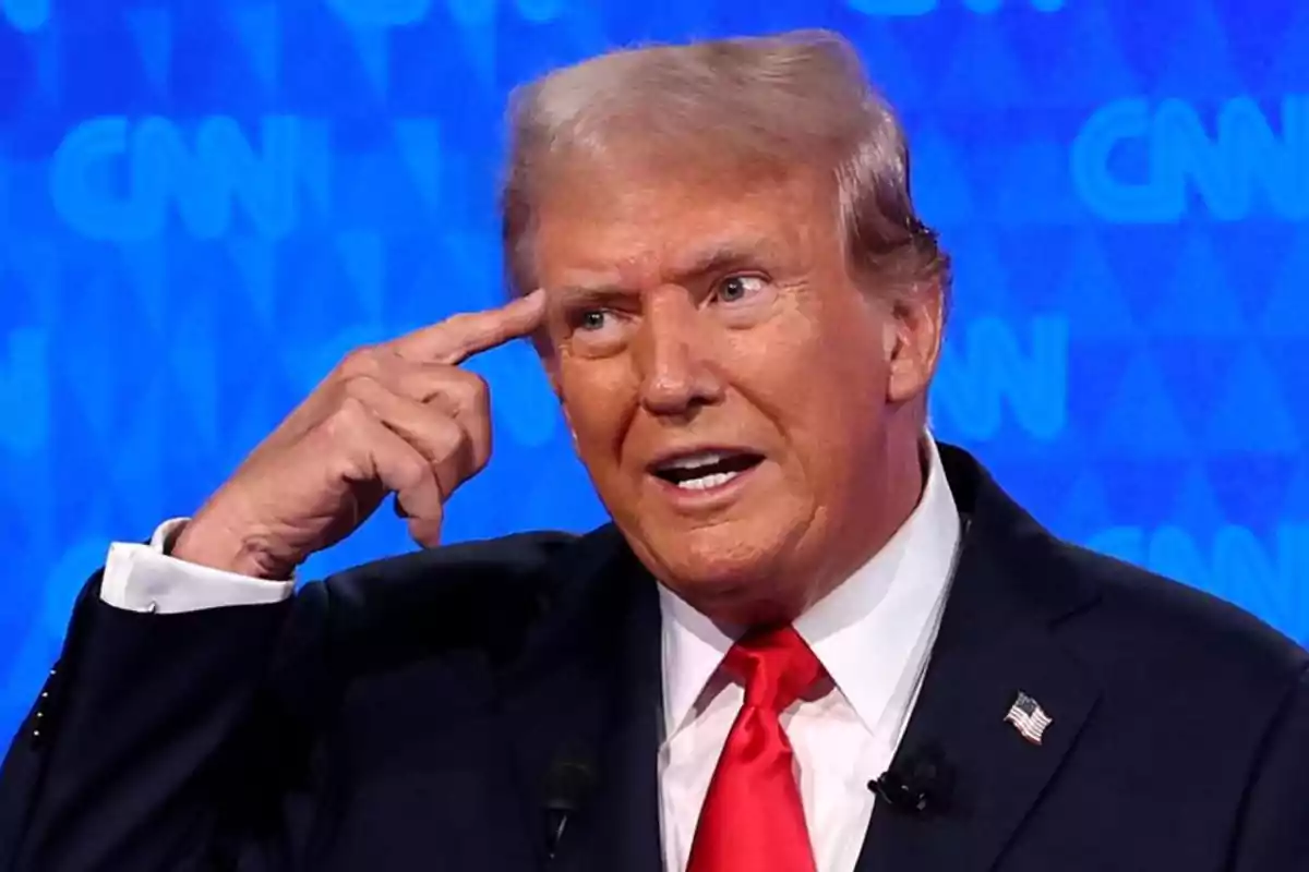 Hombre en traje oscuro con corbata roja, señalando su sien con el dedo índice, frente a un fondo azul con logotipos de CNN.