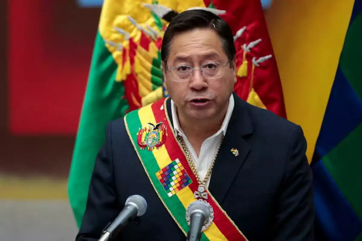 A man with glasses and a presidential sash speaks in front of two microphones with flags in the background. A man with glasses and a presidential sash speaks in front of two microphones with flags in the background.