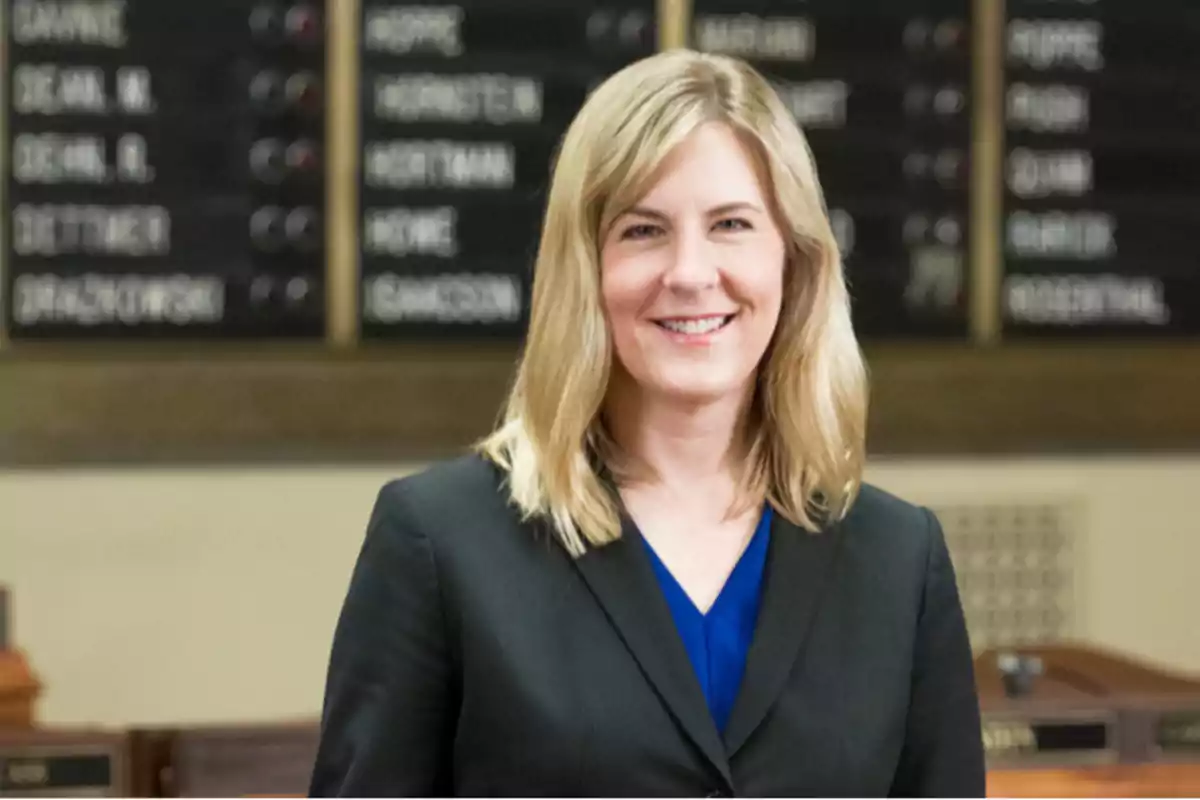Blonde woman wearing a black blazer and blue blouse smiling in a formal setting with name boards in the background Blonde woman wearing a black blazer and blue blouse smiling in a formal setting with name boards in the background
