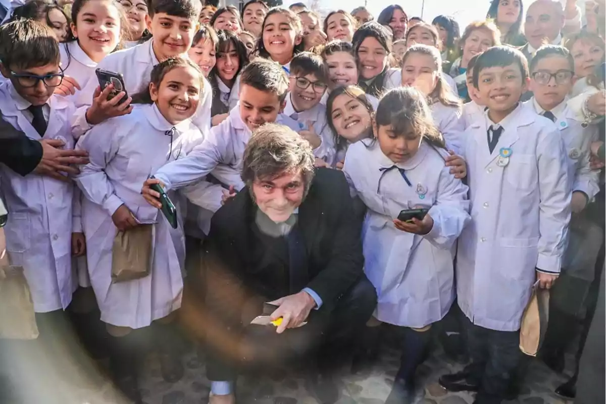 Un grupo de niños con batas blancas sonríe mientras rodea a un hombre agachado en el centro.