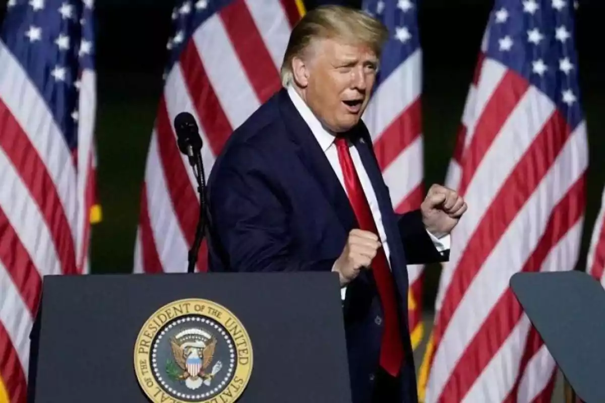 Hombre de traje oscuro y corbata roja gesticulando frente a un podio con el sello presidencial de Estados Unidos y varias banderas estadounidenses de fondo.