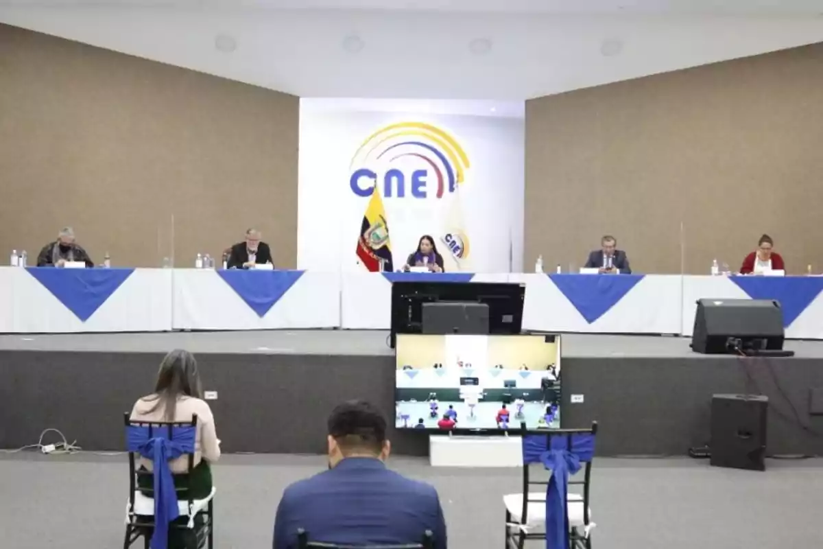 Personas sentadas en una mesa larga con manteles blancos y azules en un auditorio con el logo del CNE en la pared y una pantalla en el centro.