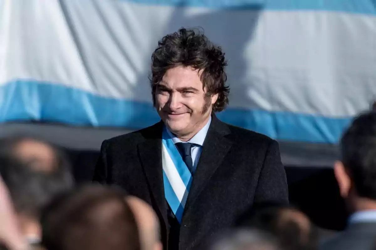 Un hombre con traje oscuro y corbata azul claro sonríe frente a una bandera con franjas celestes y blancas.