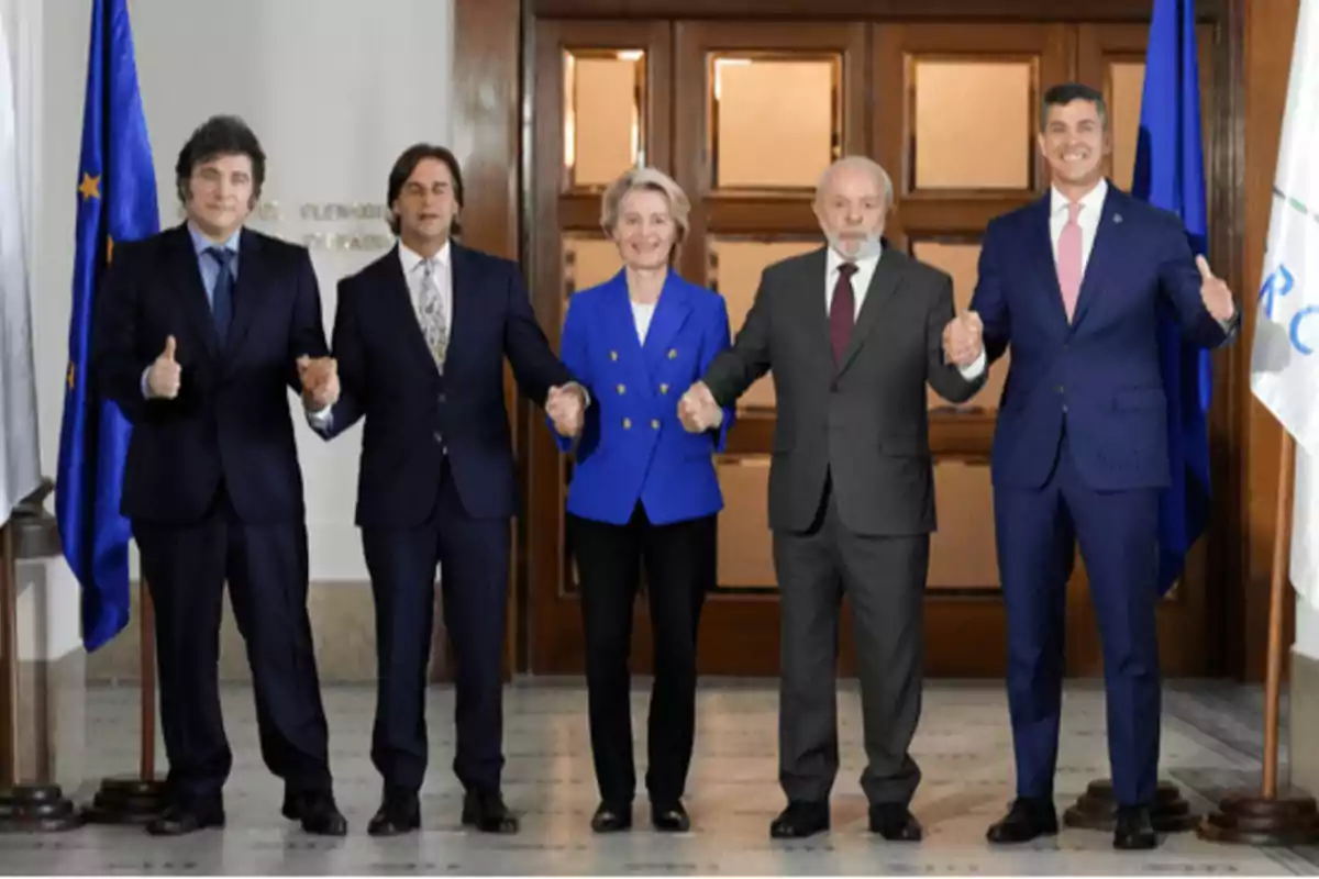 Cinco personas de pie, vestidas formalmente, posan frente a una puerta de madera, sosteniéndose de las manos y sonriendo, con banderas a los lados.