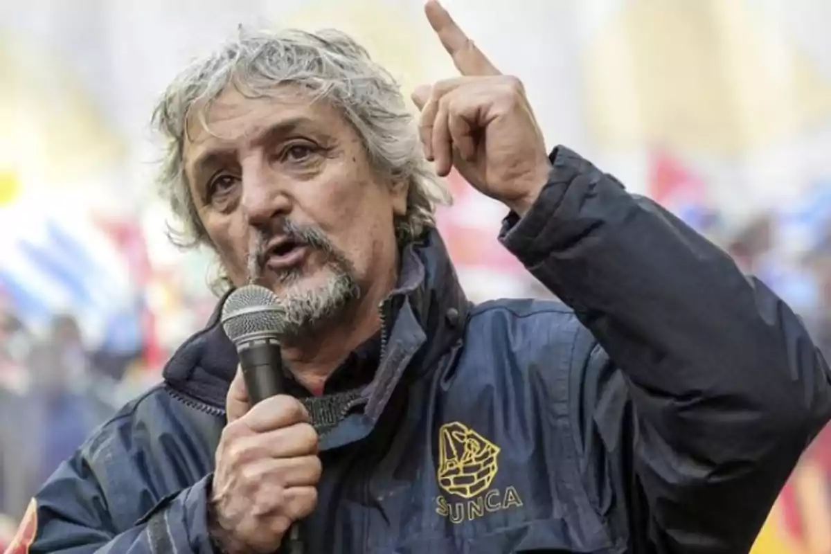 Gray-haired, bearded man speaking into a microphone and raising a finger while wearing a blue jacket with the SUNCA logo at an outdoor event Gray-haired, bearded man speaking into a microphone and raising a finger while wearing a blue jacket with the SUNCA logo at an outdoor event