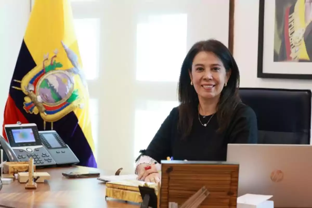 Mujer sentada en un escritorio de oficina con una bandera de Ecuador al fondo y una computadora portátil frente a ella