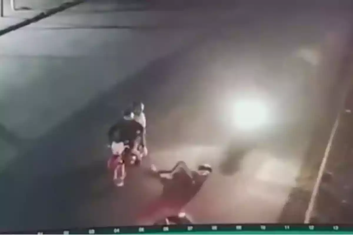 Dos personas en una motocicleta en una calle oscura, con una persona en el suelo frente a ellos.