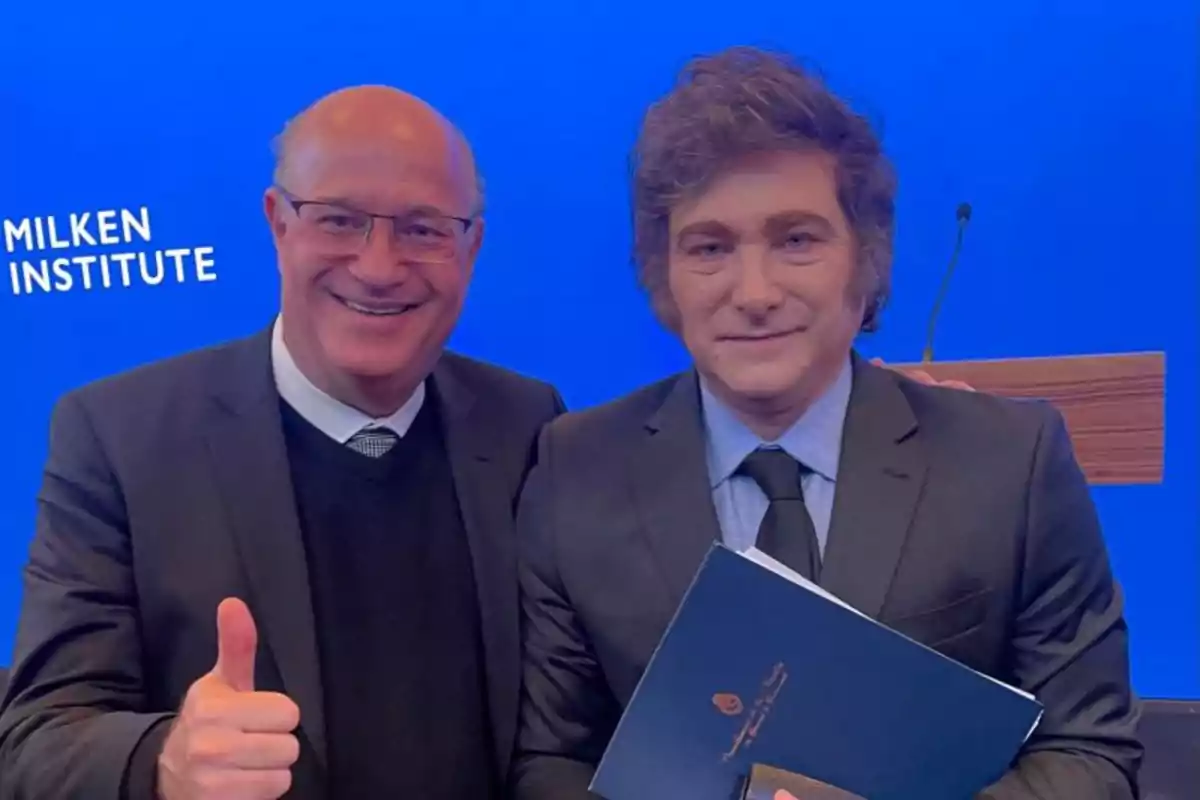 Two men pose together in front of a blue background with the text "Milken Institute," one of them holding a document and the other giving a thumbs-up.