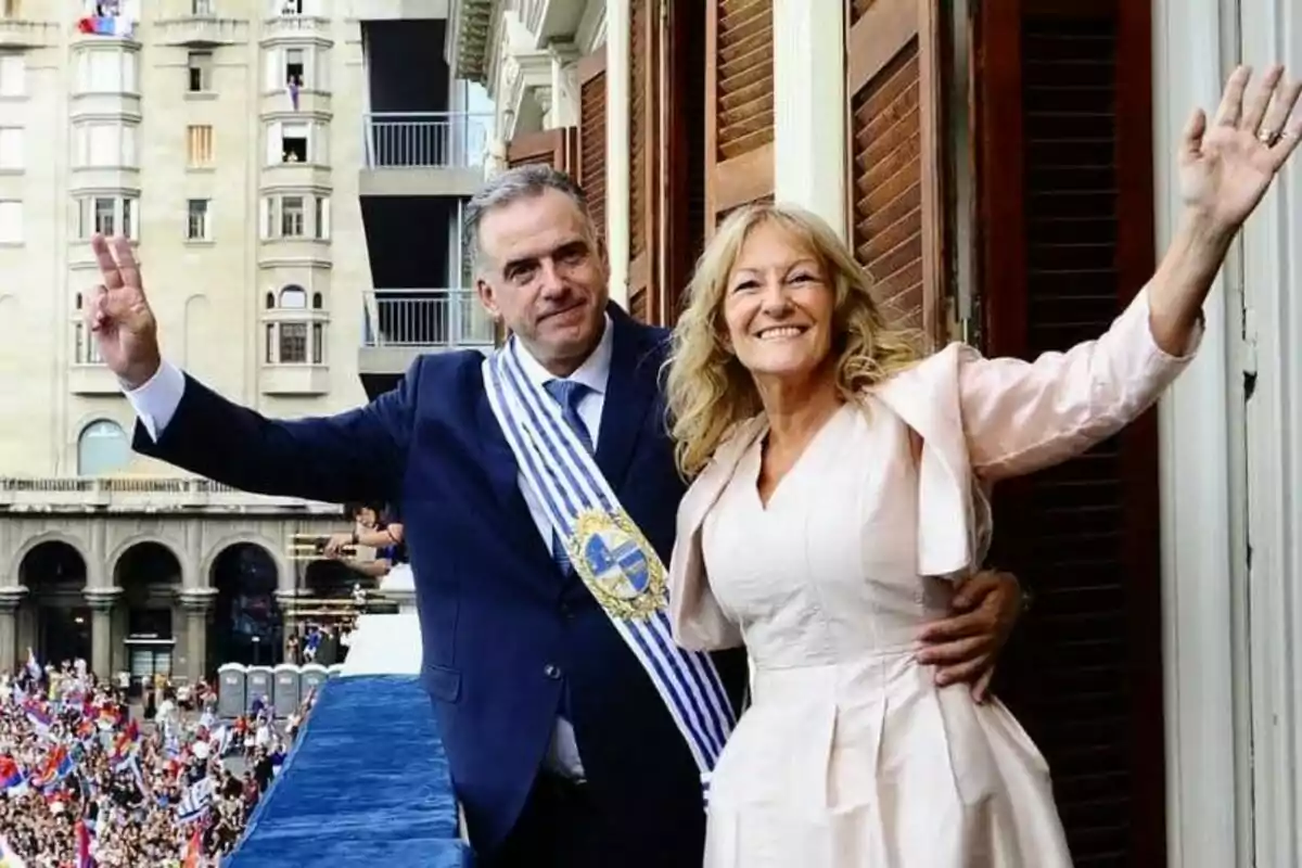 Una pareja sonriente saluda desde un balcón, con una multitud y edificios de fondo.