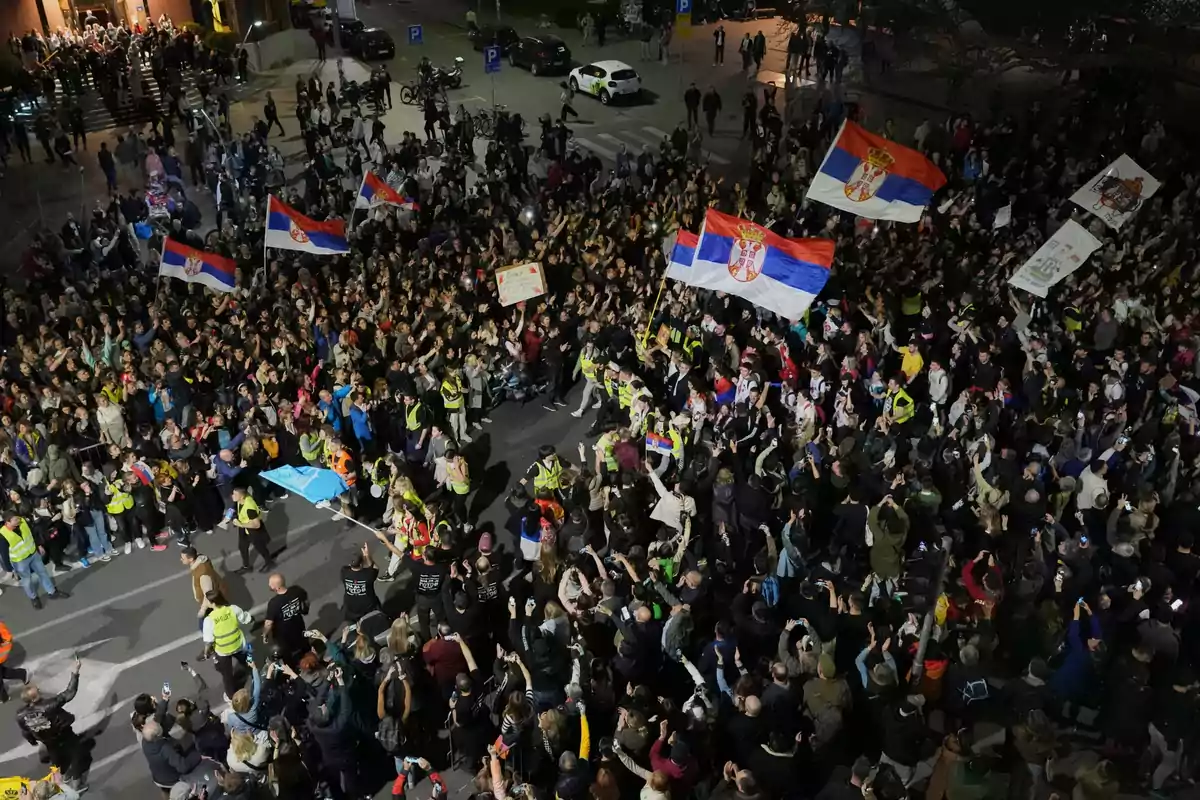 Una multitud de personas se reúne en una manifestación nocturna, ondeando banderas de Serbia y sosteniendo carteles, mientras la policía vigila el evento.