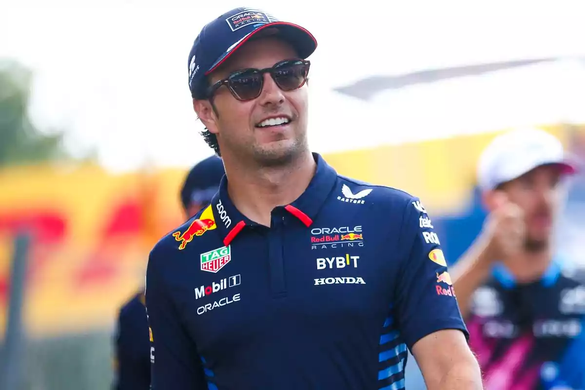 A man with sunglasses and a cap is wearing a racing team t-jersey with several logos.