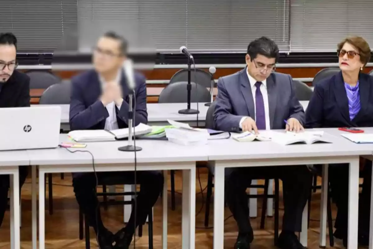 Cuatro personas sentadas en una mesa de oficina con documentos y computadoras, dos de ellas revisando papeles y una usando una laptop Cuatro personas sentadas en una mesa de oficina con documentos y computadoras, dos de ellas revisando papeles y una usando una laptop