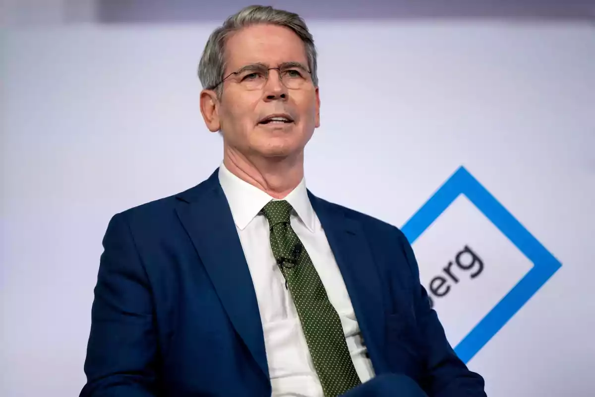Un hombre con traje azul y corbata verde habla en un evento con un logotipo de fondo. Un hombre con traje azul y corbata verde habla en un evento con un logotipo de fondo.