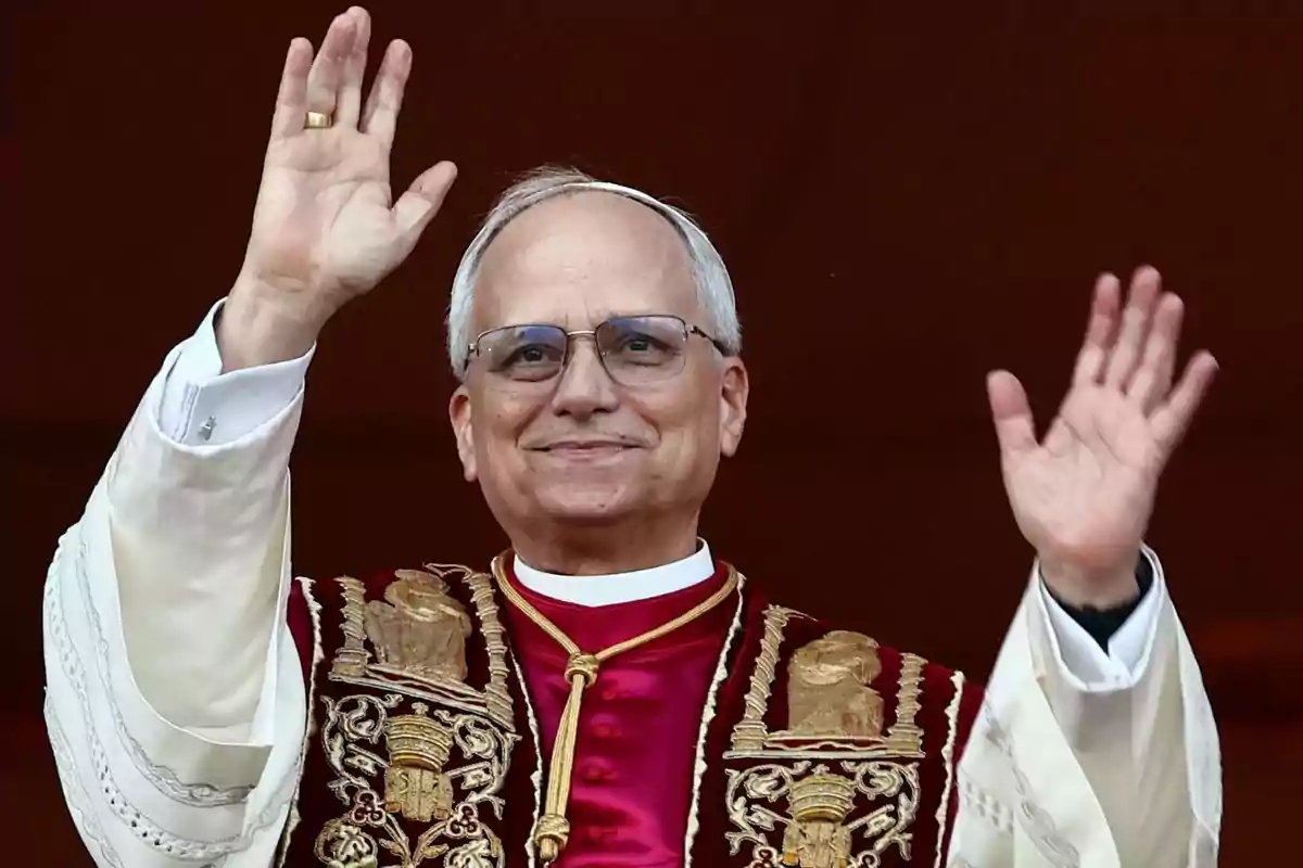 Un hombre con vestimenta religiosa y gafas sonríe mientras levanta ambas manos en un gesto de saludo. Un hombre con vestimenta religiosa y gafas sonríe mientras levanta ambas manos en un gesto de saludo.