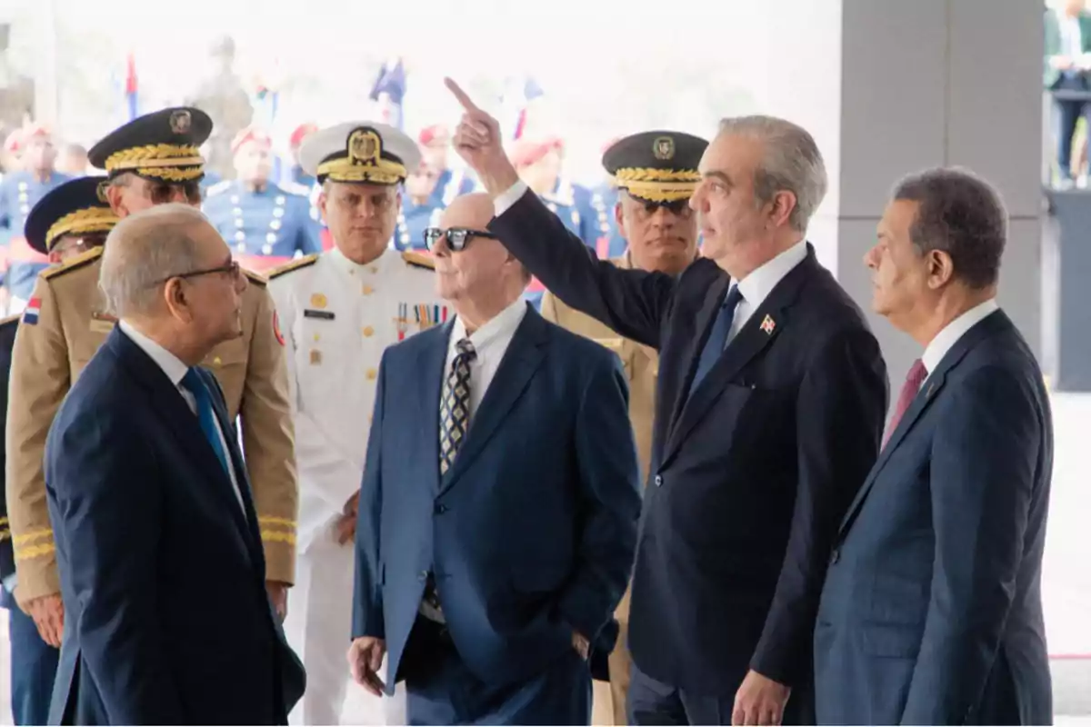 Grupo de hombres vestidos de traje y militares uniformados en un evento oficial uno de ellos señala hacia arriba mientras los demás observan atentamente