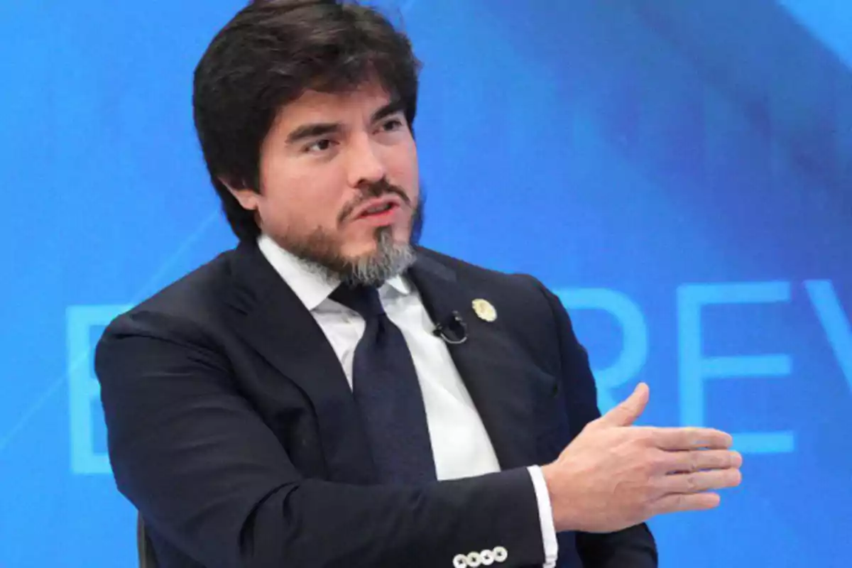 Un hombre con barba y traje oscuro gesticula mientras habla en un entorno profesional con fondo azul.