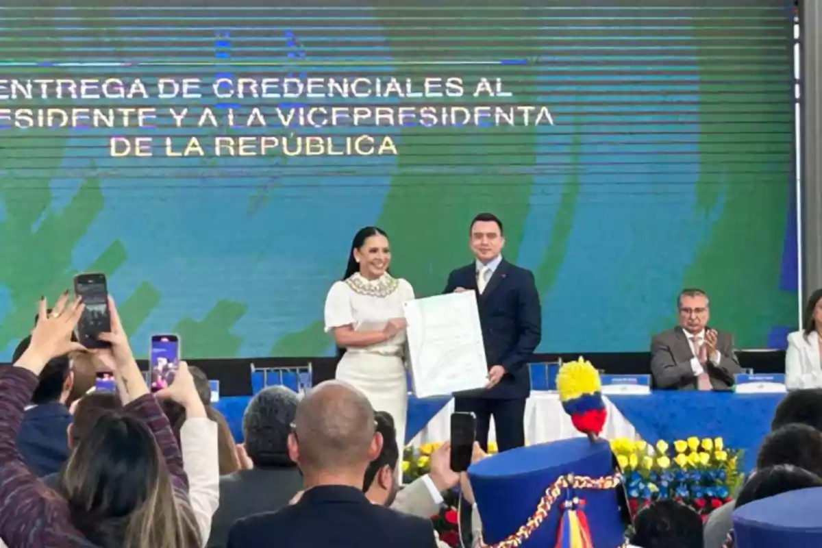 Una mujer y un hombre sostienen un documento grande en un evento formal, mientras varias personas en el público toman fotos con sus teléfonos.