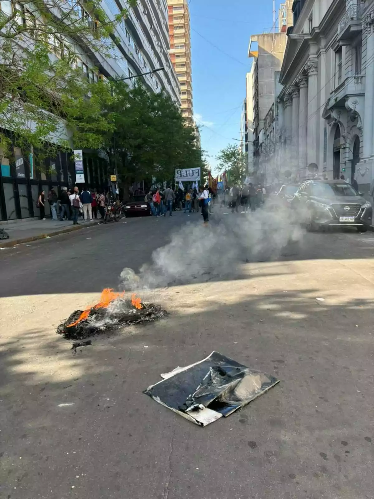 Una calle con un pequeño fuego en el suelo y humo, mientras un grupo de personas se reúne al fondo con pancartas.