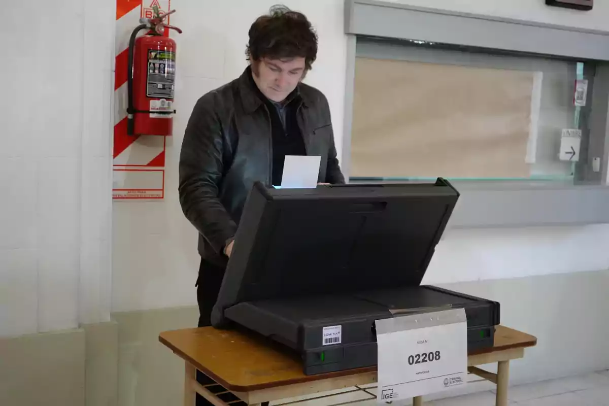 Un hombre emite su voto en una máquina de votación electrónica en un centro de votación, con un extintor de incendios visible en la pared detrás de él. Un hombre emite su voto en una máquina de votación electrónica en un centro de votación, con un extintor de incendios visible en la pared detrás de él.