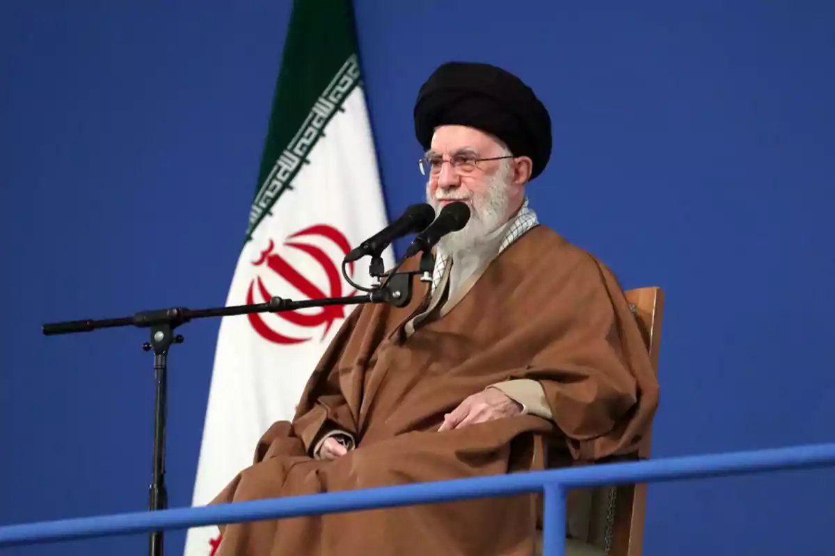 Elderly man with a beard and a black turban speaking into a microphone in front of an Iranian flag against a blue background