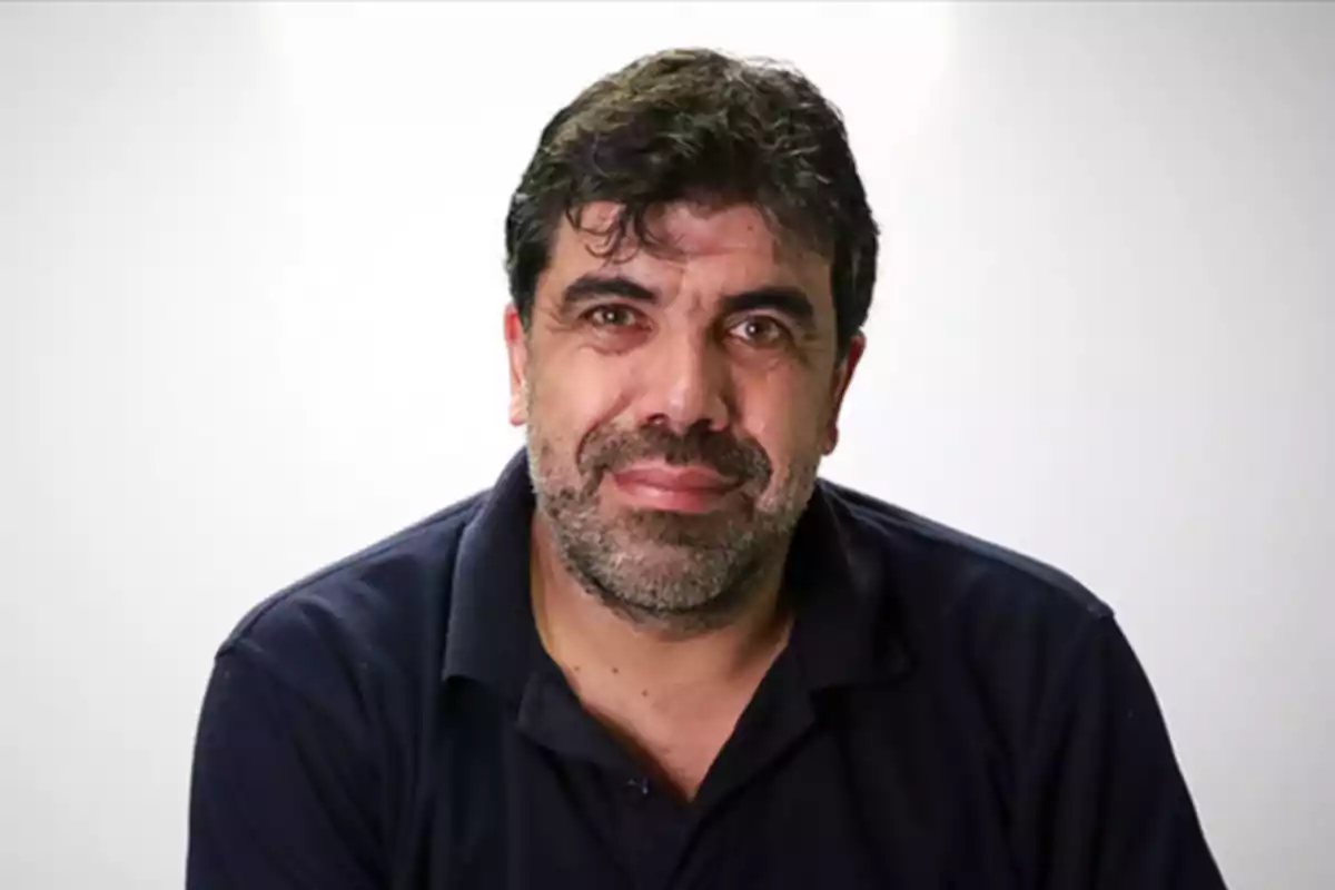 A man with a beard and dark hair is wearing a black jersey and looking at the camera with a neutral expression against a light background.