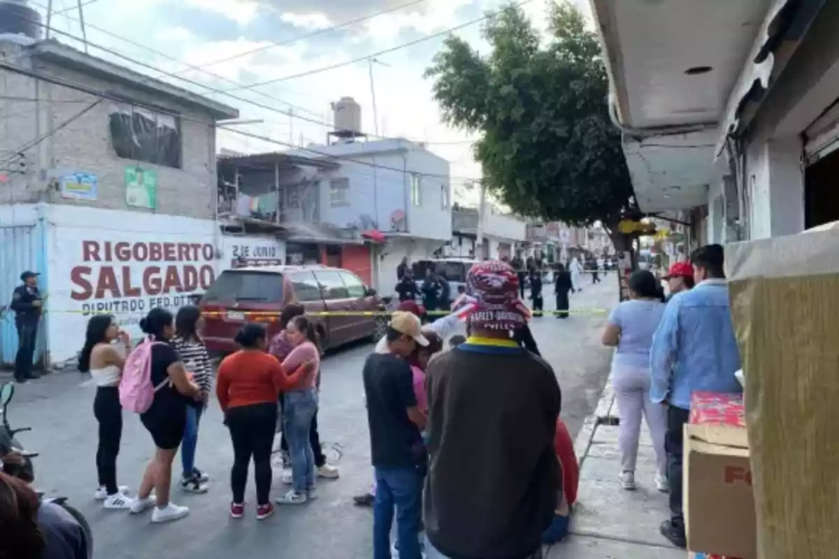 Un grupo de personas se reúne en una calle donde hay una cinta de seguridad y presencia policial en el fondo.