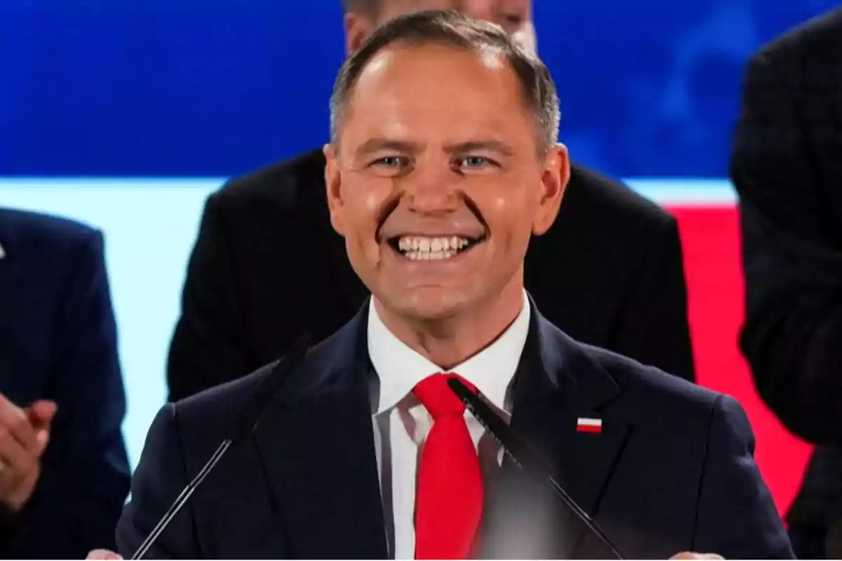 Hombre sonriente con traje y corbata roja hablando frente a micrófonos en un evento con fondo de colores azul, blanco y rojo