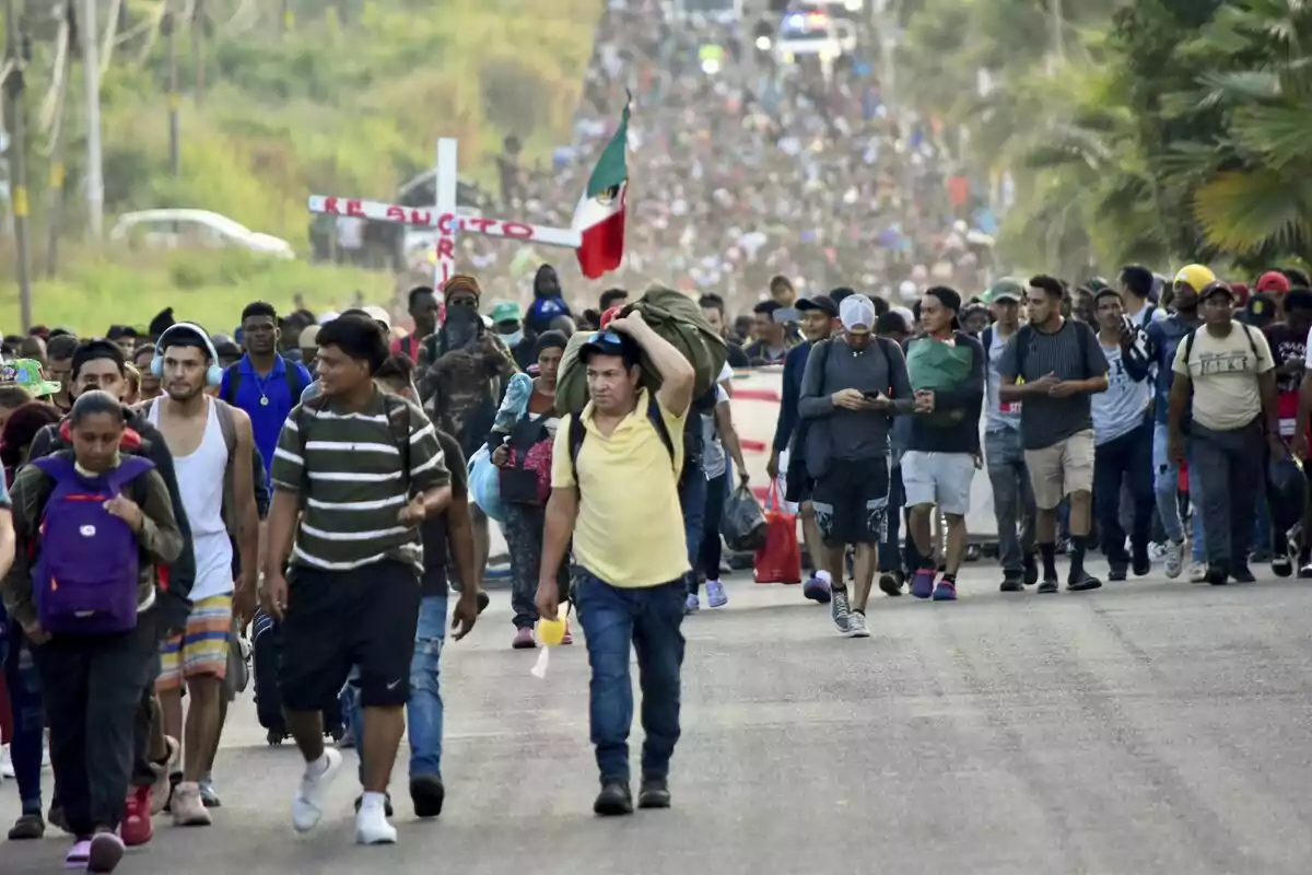 Un grupo grande de personas camina por una carretera, algunas llevan mochilas y una bandera de México.