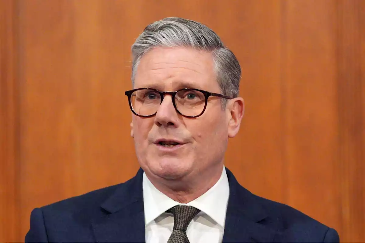 A man with glasses and gray hair is wearing a dark suit and tie in front of a wooden background. A man with glasses and gray hair is wearing a dark suit and tie in front of a wooden background.