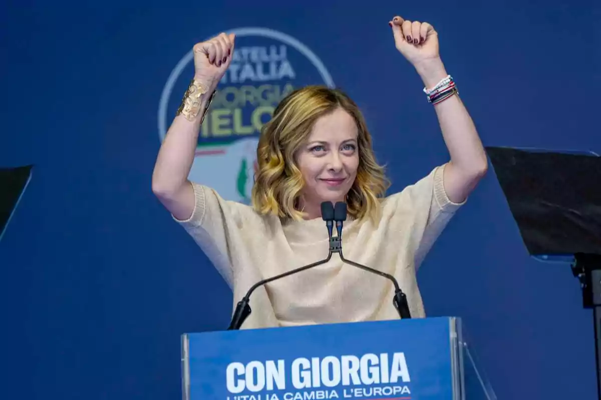 Blonde woman with raised arms smiles behind a blue podium at a political event