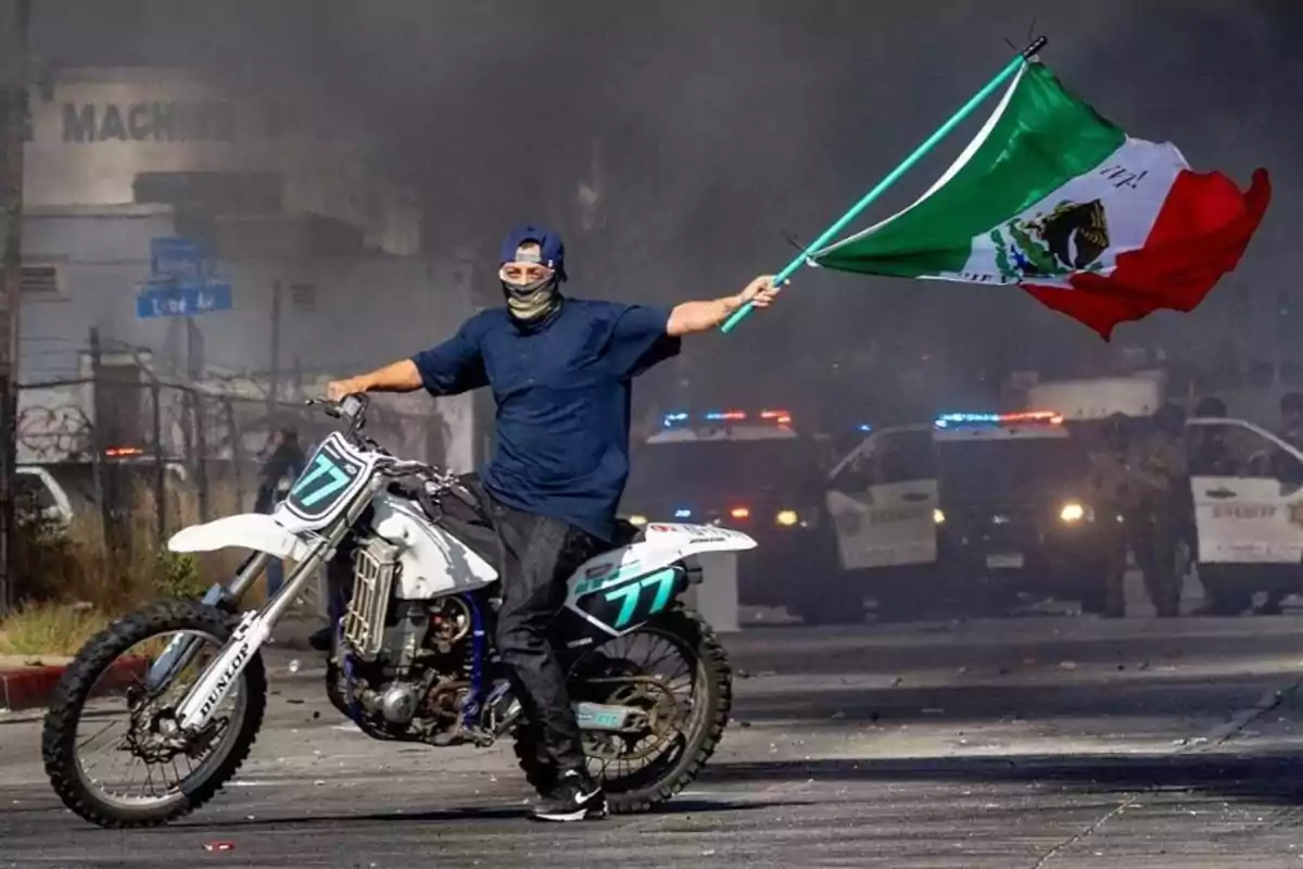 Man wearing a cap and face mask riding a motorcycle, holding a Mexican flag, with police cars and smoke visible in the background.