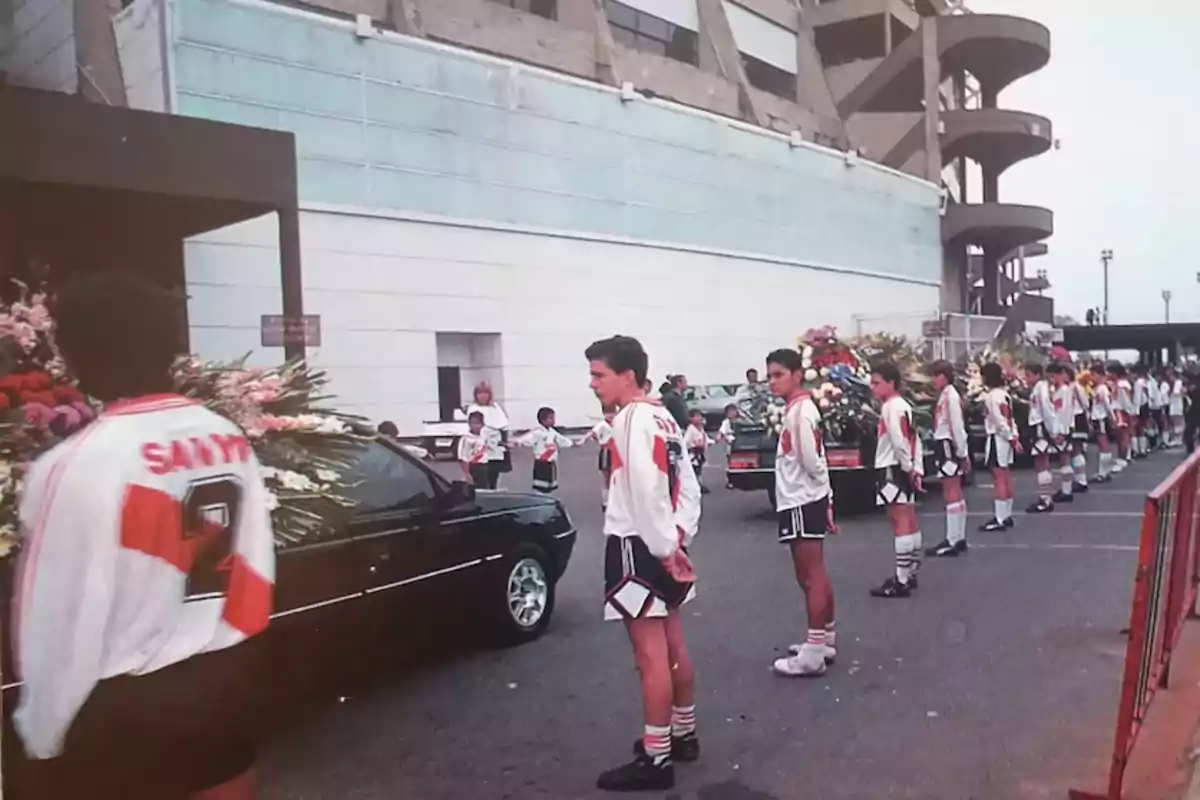Un grupo de jóvenes con uniformes deportivos forma una fila junto a un coche fúnebre adornado con flores frente a un edificio grande. Un grupo de jóvenes con uniformes deportivos forma una fila junto a un coche fúnebre adornado con flores frente a un edificio grande.