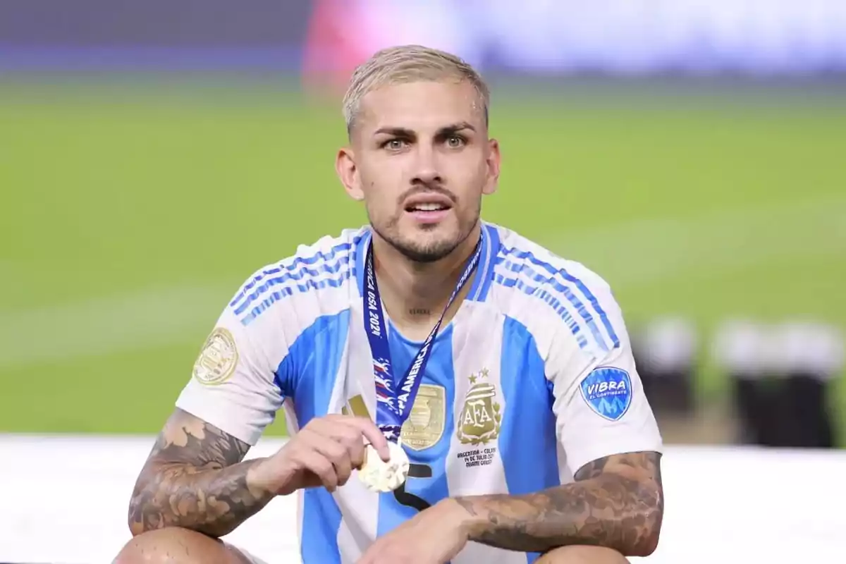 Un jugador de fútbol con uniforme de Argentina sostiene una medalla mientras está sentado en el campo.